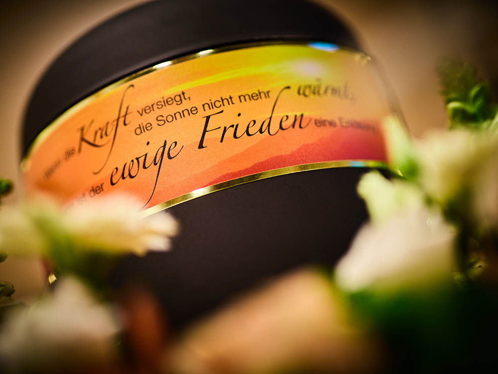 Was tun im Trauerfall: Urne im Blumenarrangement – Schwarz & Sohn Nahaufnahme einer Urne mit goldfarbener Banderole und Blüten im Vordergrund; warmes Licht, symbolische Darstellung für den Trauerfall.