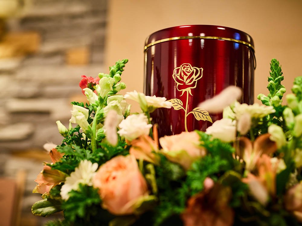 Urne mit Rosenmotiv im Blumenarrangement – Trauerhalle Versmold Rote Urne mit goldener Rosenverzierung inmitten eines Blumenarrangements; unscharfer Steinwand-Hintergrund in der Trauerhalle von Schwarz & Sohn.