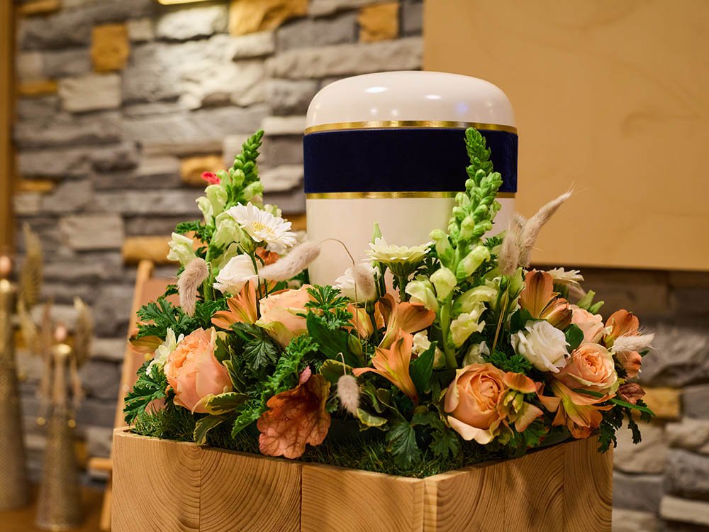 Urne mit Blumenarrangement – Trauerhalle Schwarz & Sohn Versmold Weiße Urne mit blau-goldenem Band auf Holzpodest, umgeben von Pfirsich- und Weißtönen im Blumenkranz vor Steinwand – Trauerhalle in Versmold.