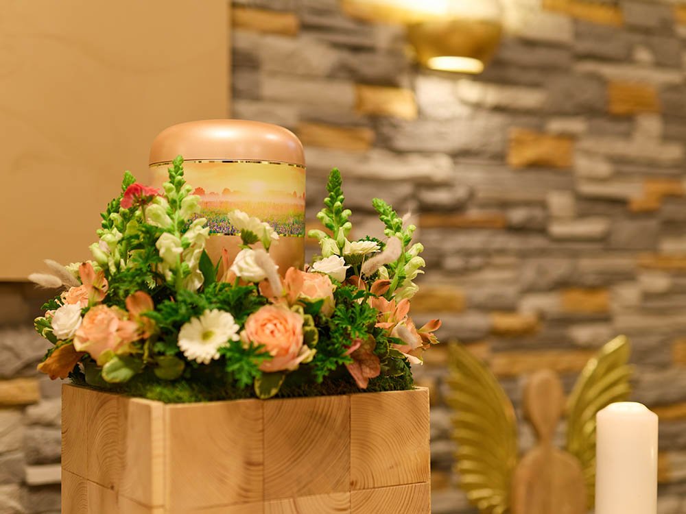Urne mit Landschaftsmotiv im Blumenarrangement – Trauerhalle Versmold Salmonfarbene Urne mit Landschaftsband auf Holzpodest, umgeben von rosa und weißen Blumen vor Steinwand in der Trauerhalle von Schwarz & Sohn.