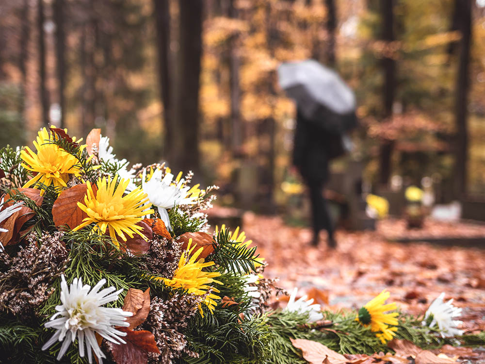 Bestattungsarten: Blumen am Grab im Herbst – Schwarz & Sohn Herbstliches Blumengesteck mit gelben und weißen Chrysanthemen im Vordergrund; unscharfe trauernde Person mit Schirm auf dem Friedhof.