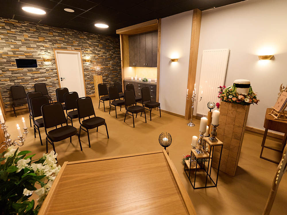 Trauerhalle Versmold – Blick vom Rednerpult auf Sitzreihen Trauerhalle von Schwarz & Sohn in Versmold mit Bestuhlung, Urnenpodest mit Blumen und Kerzenständern; Blick vom Rednerpult in den Saal.