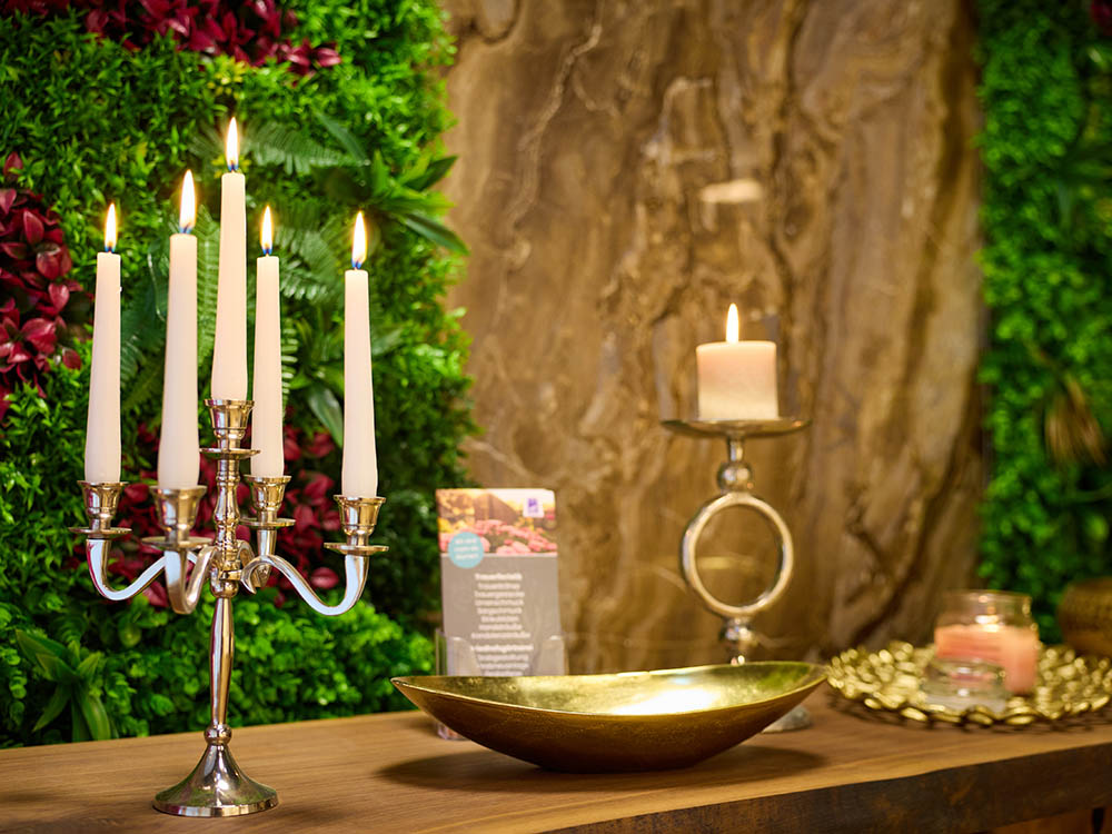 Bestattungsvorsorge – Kerzenleuchter & Kerze vor Grünwand Silberner Kerzenleuchter mit fünf brennenden Kerzen, einzelne Stumpenkerze und goldene Schale auf Holztisch vor Grünwand – Symbolfoto zur Bestattungsvorsorge.